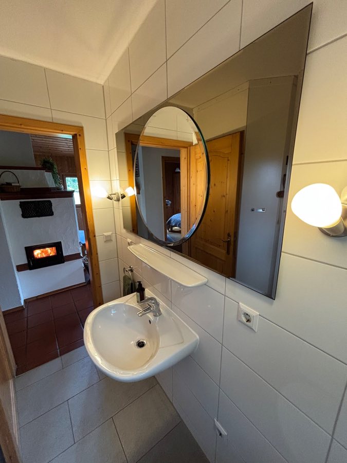 Gefliestes Badezimmer mit Waschbecken, großem Spiegel und Blick durch die Tür zum Kachelofen