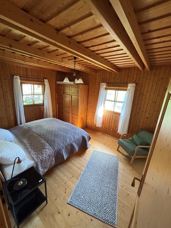 Helles Schlafzimmer mit Doppelbett, sichtbaren Deckenbalken, Kleiderschrank und grünem Sessel
