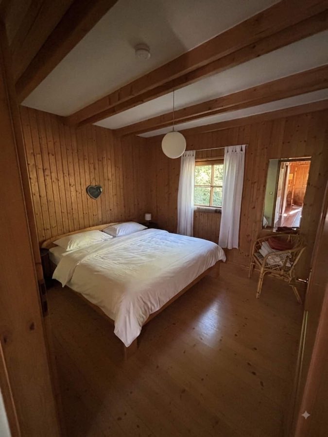 Schlafzimmer mit holzverkleideten Wänden, Doppelbett mit weißer Decke und Korbstuhl am Spiegel