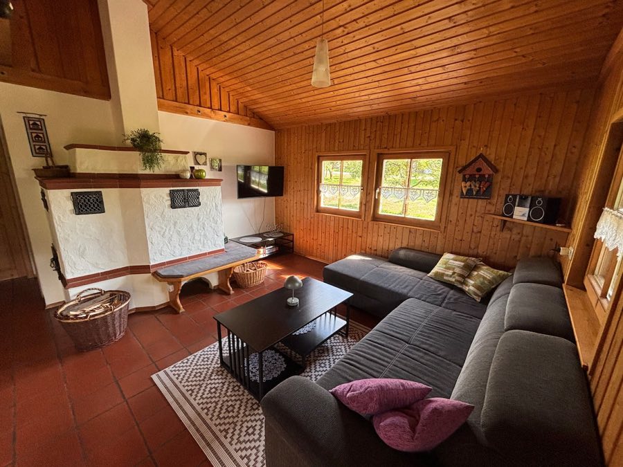 Wohnzimmer mit großer grauer Eckcouch, Holzdecke, Fernseher und weißem Kachelofen